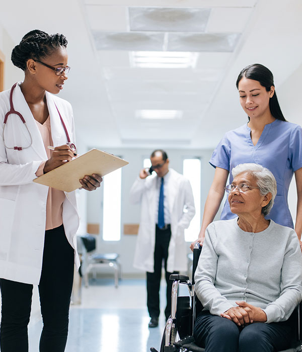 Female doctor with clipboard facing female patient in wheelchair with female nurse pushing