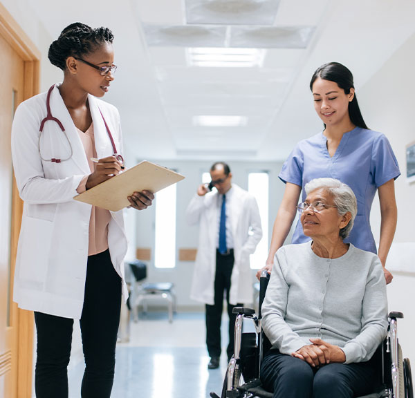 Hospital hallway with female doctor speaking to an elderly female patient in a wheelchair being pushed by a female nurse