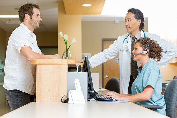 Male patient leaning on counter in waiting area as he chats with male doctor and female receptionist on the other side